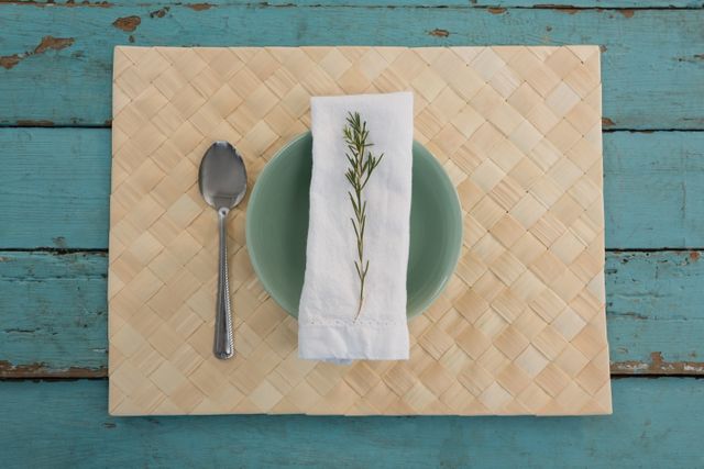 Overhead view of a rustic table setting featuring a woven placemat, a teal plate, a white napkin with a sprig of herb, and a spoon on weathered wooden planks. Ideal for use in articles or advertisements about dining, home decor, rustic themes, or farmhouse style. Perfect for blogs, social media posts, or websites focusing on lifestyle, food, and interior design.