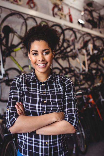 Young female mechanic standing confidently with arms crossed in a bicycle workshop. Ideal for use in articles or advertisements related to bicycle repair services, professional workshops, or promoting skilled trades. Can also be used in content focusing on women in traditionally male-dominated professions, showcasing confidence and expertise.