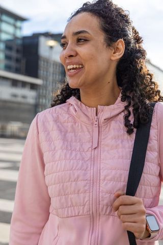 Young woman smiling while walking across urban crosswalk wearing pink jacket and smartwatch