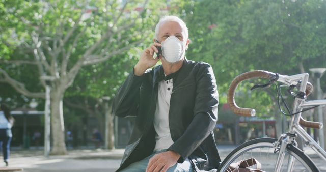 Senior man wearing mask talking on phone sitting on bench next to bicycle in urban park with trees. Ideal for concepts of social distancing, outdoor activities, senior health, communication, and pandemic safety. Perfect for articles, advertisements, blogs related to cycling, senior lifestyle, and public health messages.