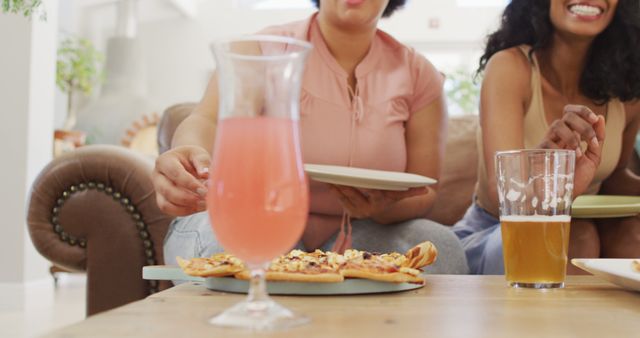 Two women sitting on a couch socializing while enjoying pizza and drinks in a cozy living room environment. The table in front of them has a pizza and different drinks including a colorful beverage and a beer. Ideal for use in contexts related to friendship, leisure time, casual dining, home gatherings, and social bonding.