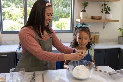 Mother and daughter baking together in bright kitchen cracking egg into glass mixing bowl