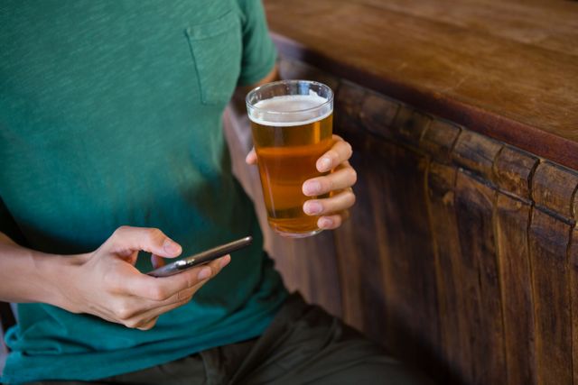 Man holding a glass of beer in one hand and using a smartphone with the other at a bar. Ideal for themes related to casual lifestyle, socializing, technology use in everyday life, and leisure activities. Suitable for advertisements, blog posts, or articles about modern social habits, bars, and pubs.
