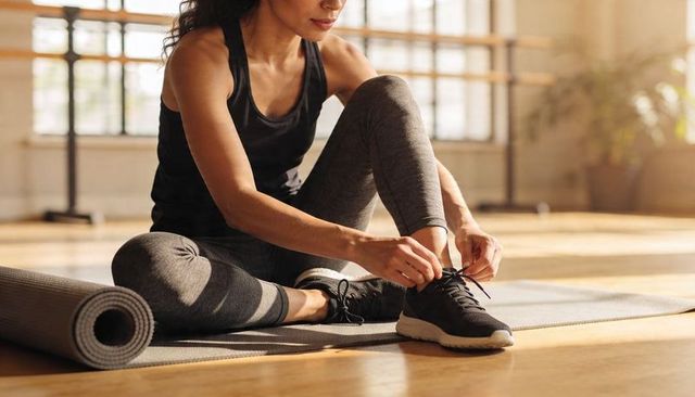 Woman tying running shoe on yoga mat in sunlit studio during morning workout
