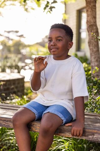 African american boy sitting on wooden bench in sunlit garden smiling and waving