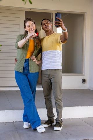 Diverse couple taking selfie on front porch holding smartphone and red key fob