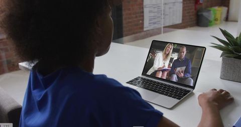 Woman joining remote video meeting on laptop at open-plan coworking desk with succulent and brick wa