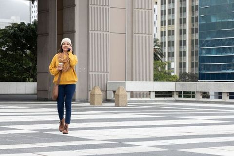 Woman walking and talking on phone holding coffee in urban plaza with modern buildings