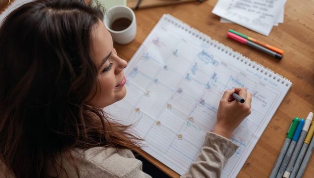 Young woman planning monthly calendar at home desk, writing in spiral planner with colorful markers