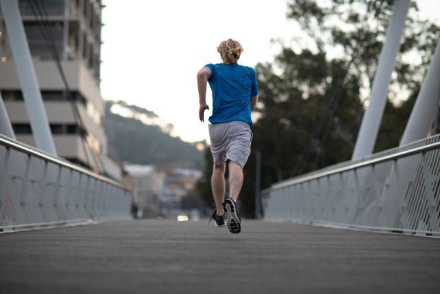Rear view of a fit Caucasian man with long blonde hair wearing sportswear exercising outdoors in the city on a sunny day with blue sky, running on a footbridge.