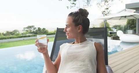 Relaxing woman reclining by infinity pool holding lemon water, sunglasses on head