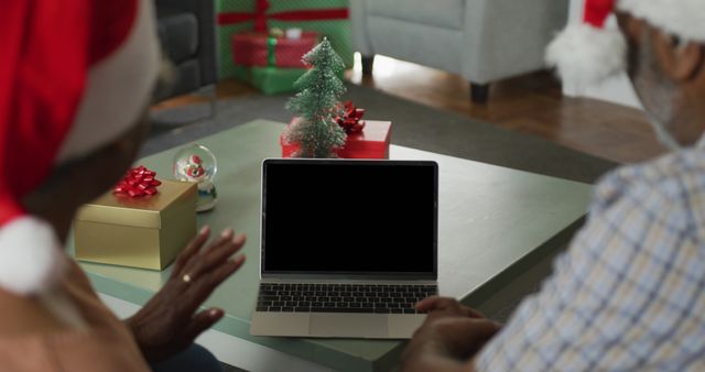 Elderly couple wearing Santa hats video calling family on laptop during Christmas. Wrapped gifts and festive decorations on table add to the holiday ambiance. Perfect for themes such as holiday season, virtual gatherings, family connections, and Christmas celebrations.