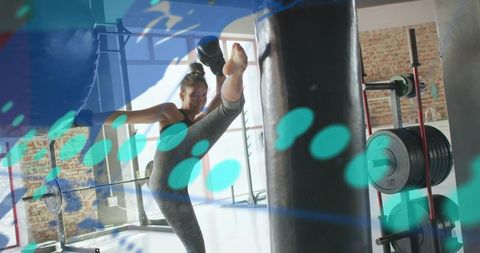 Female athlete kicking high in gym training with punching bag, weights and focus