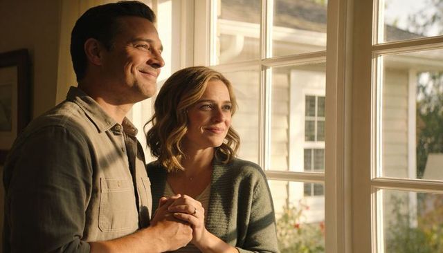 Sunlit mature couple holding hands, gazing out window with engagement ring visible