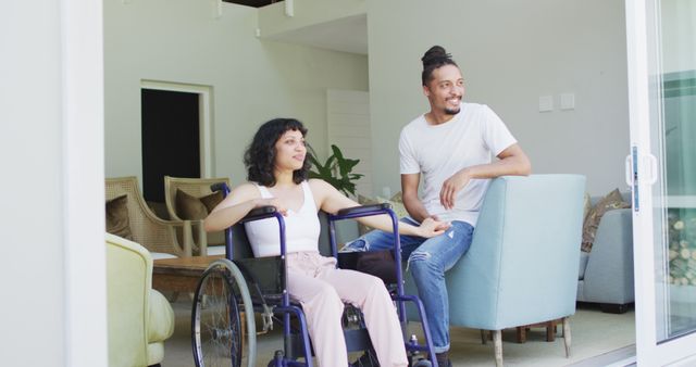 Happy biracial woman in wheelchair and smiling male partner looking out of terrace in living room. wellbeing and domestic lifestyle with physical disability.