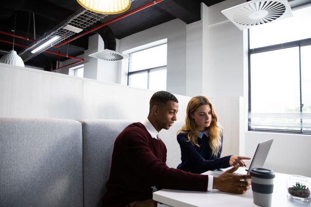 Young African American man and Caucasian woman collaborating on a laptop in a modern office. Ideal for illustrating teamwork, diversity in the workplace, modern business environments, and professional collaboration.