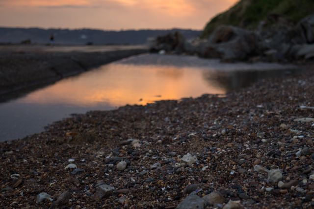 Coastal sunset over a peaceful pebble beach with calm water reflecting the twilight sky. Ideal for nature scenes, relaxation themes, travel advertisements, and coastal artwork backdrops.