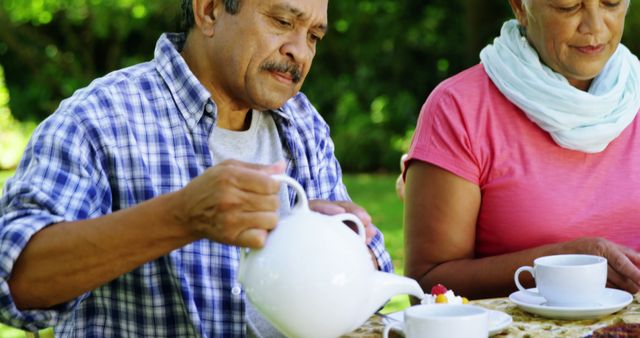 A senior Asian man is pouring tea from a white teapot for a senior Asian woman during an outdoor tea time, with copy space. They appear to be enjoying a peaceful moment together in a garden setting.