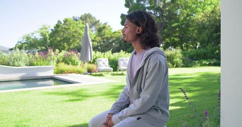African american man sitting by pool in gray hoodie reflecting in sunlit backyard garden
