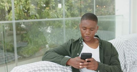 African american man using smartphone on sofa by glass window with lush patio greenery