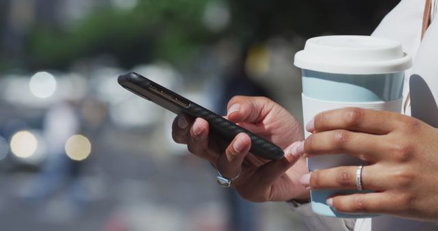This image captures a person holding a smartphone and a coffee cup outdoors. The scene suggests a casual urban setting and highlights the use of modern technology for communication or leisure while on the go. Ideal for illustrating themes of technology in everyday life, urban living, coffee culture, or mobile communication.