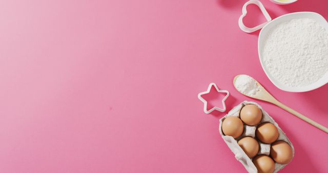Eggs in a carton, a bowl of flour, a wooden spoon, and cookie cutters including star and heart shapes arranged on a bright pink background. Perfect for illustrating baking recipes, cooking blogs, kitchen-themed projects, or advertisements related to culinary products.