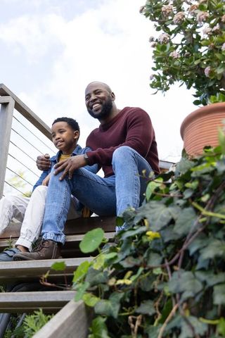 African american father and son smiling on wooden deck stairs with ivy planter