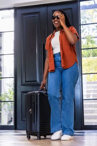 Smiling african american traveler holding suitcase, adjusting sunglasses in modern entryway