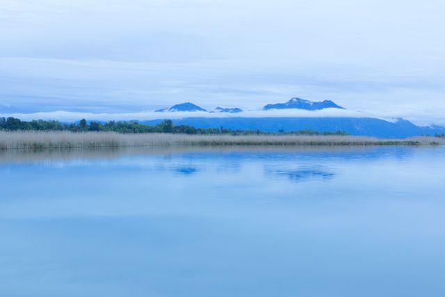 Beautiful tranquil mountain lake under a cloudy sky, surrounded by tall grass and distant mountains partially blanketed by mist. Ideal for travel blog, nature magazine, desktop wallpapers, and relaxation apps.