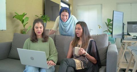 Multicultural female colleagues collaborating on laptop in modern office lounge with coffee