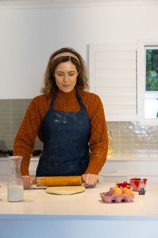 Mid-adult woman rolling dough on kitchen island wearing denim apron and cozy sweater