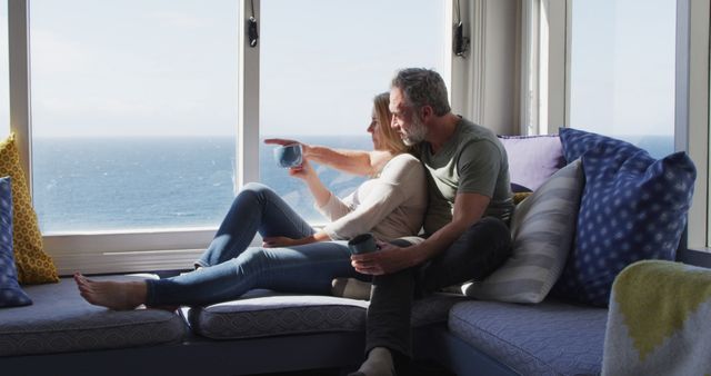 Middle-aged couple sitting on a cozy window seat, drinking coffee, looking at ocean view. Ideal for themes of romance, relaxation, vacations, serene moments, and lifestyle settings. Great for use in advertisements, travel promotions, health and wellness content, or relationship marketing.