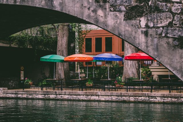 Colorful umbrellas line an outdoor dining area by a river, seen under a stone bridge. The umbrellas provide a splash of color in front of tables and chairs set up for dining. This scene can be used for depicting outdoor dining, cityscapes, urban life, or travel-related themes.