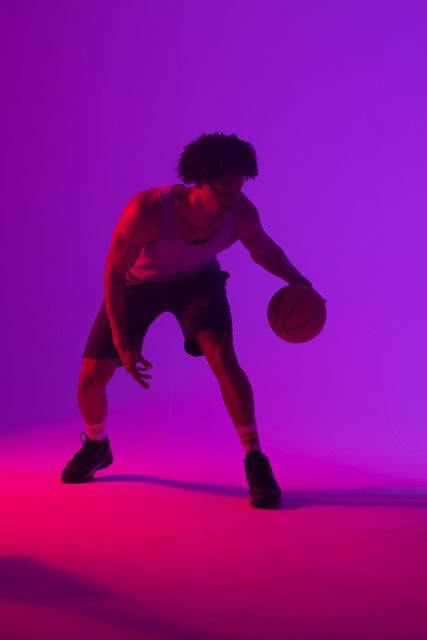 Vertical image of african american male basketball player bouncing ball, in purple light, copy space. Athlete, sport, competition, confidence and fitness concept.
