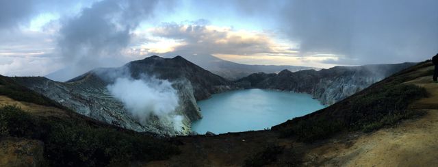 This panoramic landscape shows the famous Ijen Crater with its vivid blue acidic lake surrounded by rugged volcano walls and emitting sulfuric steam under a sky with scattered clouds. Ideal for use in travel blogs, geological studies, nature documentaries, and adventure tourism marketing.