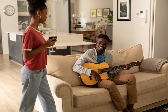 Young African American couple enjoying a relaxing evening at home. The man is playing guitar on the sofa while the woman holds a wineglass and looks at him with a smile. Perfect for themes related to love, hobbies, lifestyle, and home relaxation. Ideal for use in articles, blogs, and advertisements focusing on relationships, leisure activities, and home life.