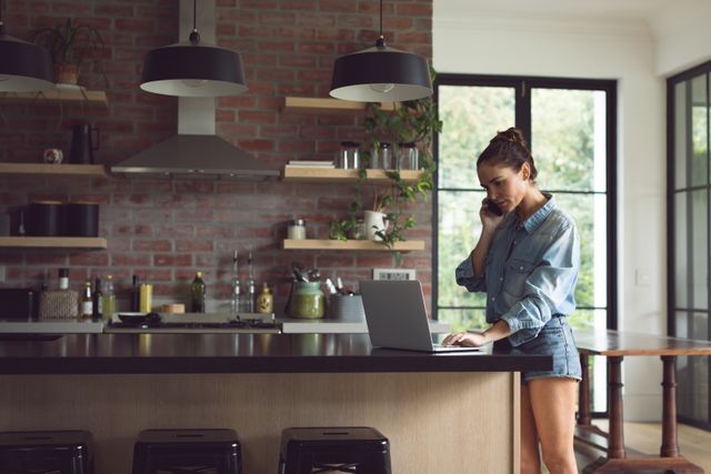 Woman multitasking by talking on phone and using laptop in a modern kitchen. Ideal for themes related to remote work, home lifestyle, technology, and contemporary living. Can be used in articles, blogs, or advertisements focusing on work-life balance, home office setups, or modern kitchen designs.