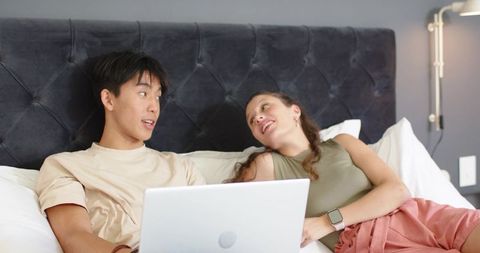 Young asian man and african american woman lounging on bed using laptop and smartwatch