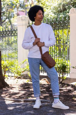 African american man standing in sunlit park holding brown crossbody bag, casual style