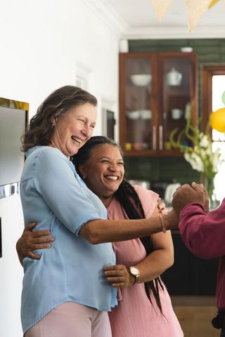 Mature women hugging and laughing during intimate family kitchen celebration with balloons
