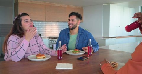 Friends enjoying casual lunch at kitchen island sharing burgers and glass soda bottles