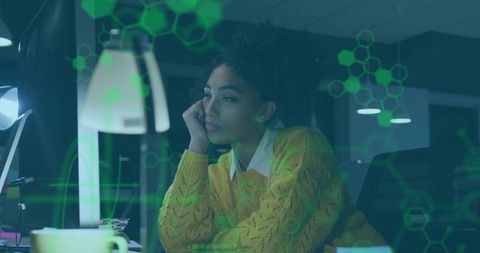 Young woman working late at desk gazing at screen with green hex technology overlay
