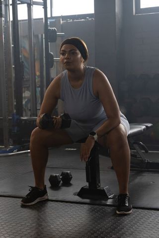 Focused african american woman strength training with dumbbell on bench in gym