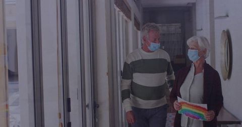 Senior couple in masks walking with rainbow painting in hallway