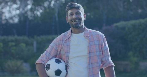 Smiling man holding soccer ball in backyard wearing pink plaid shirt and white tee