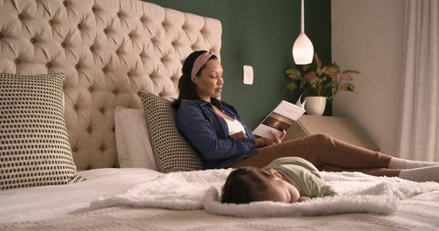 Mother reading relaxing in bedroom with sleeping infant