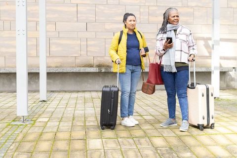 Mature women waiting at urban transit stop with rolling luggage and smartphones