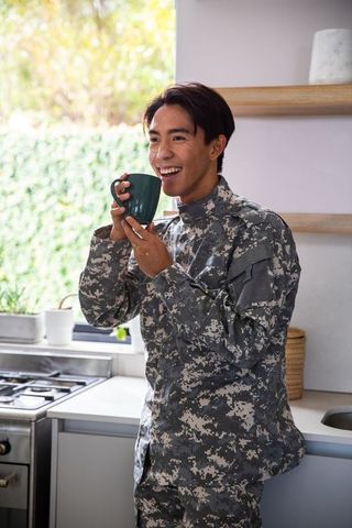 Asian service member enjoying morning coffee in sunlit minimalist kitchen, smiling