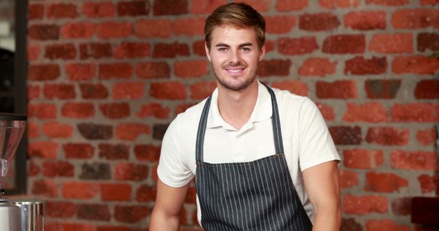 A young Caucasian male barista smiles confidently in front of a coffee machine, with copy space. His professional attire and the brick wall backdrop suggest a cozy, urban cafe environment.