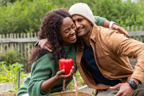 Couple embracing and laughing in backyard garden holding red bell pepper, harvesting joy
